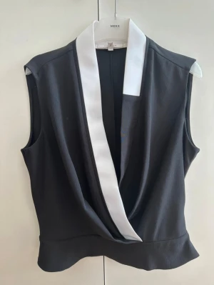 Svart omlott blus från Carven - Snygg svart blus från Carven med vit krage och omlott-detalj framtill. Ärmlös design och mjukt, lite strukturerat tyg som ger en cool och stilren look. Perfekt för dig som gillar kontraster och vill ha något som sticker ut. Storlek Fransk 42