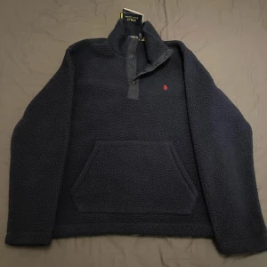Mörkblå fleece-tröja Polo Ralph Lauren - Mysig mörkblå fleece-tröja från Polo Ralph Lauren med broderad röd logga på bröstet. Tröjan har lång ärm, hög krage med knappar och en stor magficka framtill. Perfekt för att hålla dig varm under de kalla vinter dagarna.
