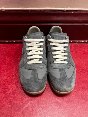 Maison Margiela sneakers  - Maison Margiela  sneakers i grå mocka och skinn med vita snören och klassisk gummisula. Snygg retrostil med rund tå och paneler i olika material. Perfekta för dig som gillar minimalistisk och stilren design.