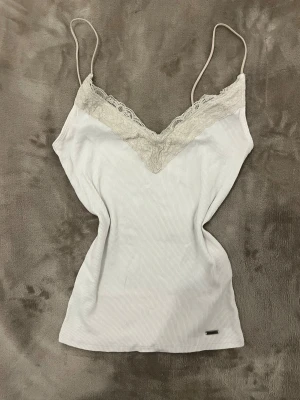 Vit ribbad linnetopp från Hollister - Supersöt vit ribbad linnetopp från Hollister med tunna justerbara axelband och spetsdetaljer vid v-ringad hals. Materialet är mjukt och stretchigt, perfekt för varma dagar. Metallbricka med Hollister-logga nedtill ger en extra touch. Bra för en y2k layered look och att ha under en henley eller babydoll top tillexempel. 