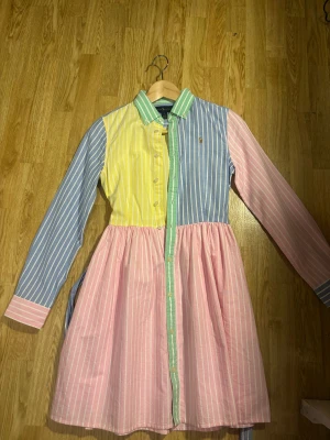 Randig skjortklänning från Ralph Lauren - Färgglad skjortklänning från Ralph Lauren i skjort-material med pastelliga ränder i blått, gult, rosa och grönt. Klänningen har lång ärm, knappar framtill, klassisk krage och avtagbart bälte i midjan. 