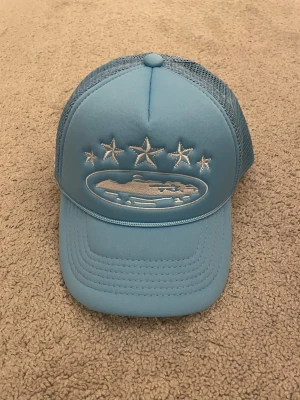 Blå Corteiz truckerkeps med mesh - Snygg ljusblå truckerkeps från Corteiz med mesh på sidorna och bak. Framtill finns broderade vita stjärnor och ett skepp, samt Corteiz-logga broderad bak. Kepsen har böjd skärm och justerbar passform. Perfekt för dig som vill sticka ut med streetwear.
