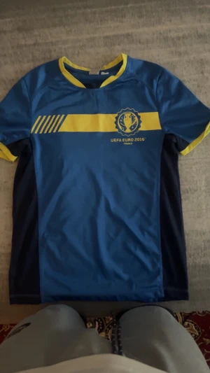 Blå UEFA Euro 2016 fotbollströja - Snygg blå fotbollströja med gula detaljer och tryck från UEFA Euro 2016. Tröjan har en gul rand över bröstet, gul krage och ärmslut. Tillverkad i lätt och ventilerande material, perfekt för match eller träning.