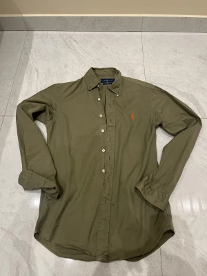 Olivgrön skjorta från Ralph Lauren - Olivgrön skjorta från Ralph Lauren med klassisk krage och knappar hela vägen. Den har lång ärm och en orange broderad logga på bröstet. Skjortan är i mjuk bomull och har en normal passform som sitter snyggt på kroppen.