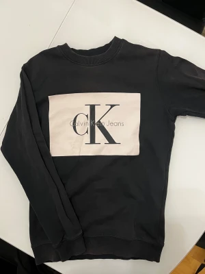 Svart sweatshirt från Calvin Klein Jeans - Svart sweatshirt från Calvin Klein Jeans med stor vit logga på bröstet. Tröjan har rund hals, långa ärmar och ribbade muddar. Materialet är mjuk bomull som känns skönt mot huden. Perfekt för en chill och stilren look.