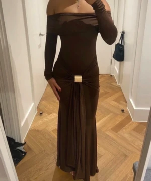 Brun offshoulder långklänning - Snygg brun långklänning med offshoulder och långa ärmar. Klänningen har en tight passform upptill och ett dekorativt knyt med guldfärgad detalj i midjan. Materialet är mjukt och följsamt, perfekt för dig som gillar en elegant och trendig look.