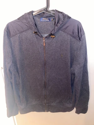 Polo Ralph Lauren Cardigan - Snygg grå cardigan från Polo Ralph Lauren med huva och snörning. Jackan har dragkedja framtill och diskret logga på bröstet. Materialmix med polyester på axlar och huva, resten i mjuk bomull. Perfekt för dig som gillar stilren och sportig look. Skicket är väldigt bra och pris diskuterbart. Kontakta mig om frågor!😊