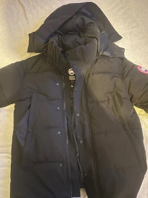Svart Canada Goose dunjacka S - Svart dunjacka från Canada Goose med klassisk patch på ärmen. Jackan har huva, dragkedja och knappar framtill samt stora fickor. Fylld med dun och fjädrar för extra värme, perfekt för kalla dagar. Snygg och stilren design som är lätt att matcha. Knapparna funkar inte bara som 4 uppe o dom 2 nere inte