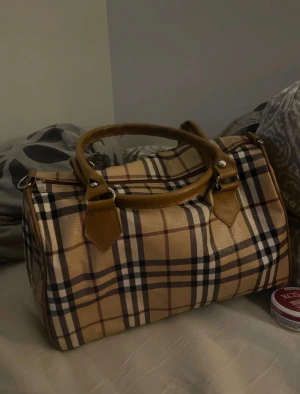 Rutig handväska med bruna detaljer - Snygg handväska i klassiskt rutig mönster med beige, bruna och svarta toner. Väskan har två stadiga handtag i brunt skinnliknande material och silverfärgade metalldetaljer. Perfekt storlek för att få plats med allt nödvändigt och har en boxig form.