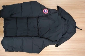 Svart pufferjacka från Canada Goose - Svart pufferjacka från Canada Goose med huva och klassisk rund logotyp på ärmen. Jackan har en stilren, quiltad design och är perfekt för kalla dagar. Fylld med dun och har en loose passform som ger en cool och avslappnad vibe.