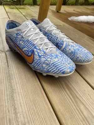 Nike Mercurial Zoom CR7 Vapor 15 Elite - Nike Mercurial Zoom fotbollsskor med grymt mönstrad design i blått och grått, med orange swoosh på sidan. Skorna har snörning, platt sula med dobbar och är tillverkade i syntetmaterial med stickad ovandel för extra komfort. Perfekt för dig som vill sticka ut på planen.