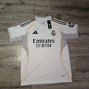 Real Madrid 25/26 M - Säljer en officiell Real Madrid fotbollströja från Adidas i storlek M. Tröjan är vit med svarta Adidas-ränder på axlarna, gula detaljer och klubbmärke på bröstet. Materialet är lätt polyester, perfekt för träning eller match. Emirates Fly Better tryck på framsidan.