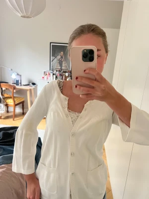 Vit kofta med knappar och fickor - Säljer en stilren vit kofta med knappar framtill och två fickor. Koftan har trekvartsärmar och en ribbad kant längs knäppningen. Perfekt att slänga över ett linne eller t-shirt för en chill look.