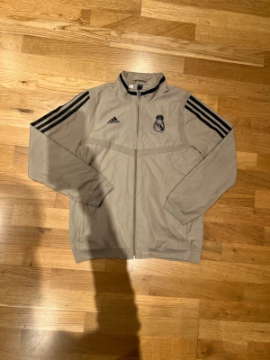 Real Madrid Tränings jacka - En tunn anthem jacka designad av adidas med Real Madrid logga. Köpt för runt 1100 från unisport. Jackan har två små fläckar som man inte tänker på om man inte letar efter defekter på den. Storleken är 152 men den passar dig som är runt 160. Hör av dig om du har någon mer fråga. Priset är prutbart via enkel affär