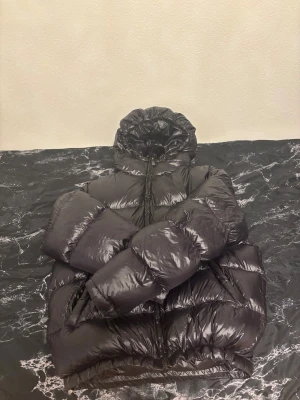 Svart pufferjacka från Zara med huva - Säljer en svart pufferjacka från Zara i storlek M. Jackan har en stor, mysig huva och är quiltad med breda kanaler. Materialet är glansigt och syntetiskt, och jackan stängs med dragkedja framtill. Perfekt för kalla dagar när du vill ha en cool och voluminös look.