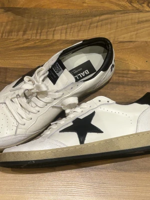 Golden Goose Ballstar sneakers vit/svart - Golden Goose Ballstar sneakers i vitt skinn med svarta detaljer och en stor svart stjärna på sidan. Skorna har snörning, rund tå och platt sula. Bak på hälen står det 'SNEAKERS' i svart text. Snygg retrostil med slitna kanter och kontrastfärger.