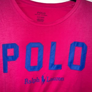 Rosa t-shirt från Polo Ralph Lauren - Snygg rosa t-shirt från Polo Ralph Lauren med stora blå bokstäver 'POLO' och logga framtill. Klassisk rund halsringning och korta ärmar. Tillverkad i mjuk bomull som känns skön mot huden. Perfekt för en avslappnad och cool stil. Har fått ett litet hål (se sista bild). ❤️Skulle passa S-L