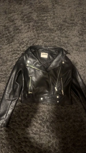 Svart skinnjacka från Noisy May - Snygg svart skinnjacka från Noisy May i klassisk bikerstil. Jackan har silvriga dragkedjor, knappar och detaljer, samt en croppad passform. Perfekt för dig som vill ha en edgy look med coola detaljer och tidlös design.