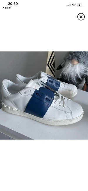 Valentino Garavani Open sneakers vit/blå - Snygga Valentino Garavani Open sneakers i vitt skinn med bred marinblå läderdetalj över snörningen. Platt sula och rund tå, samt ikoniska nitar på hälen. Perfekta för dig som gillar lyxiga och stilrena sneakers med en unik touch. Storlek 44 men passar 43 jätte bra .utan kvitto och box.
