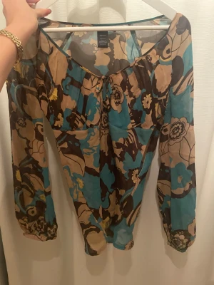 Blommig blus i silke från Filippa K - Säljer en blommig blus från Filippa K i 100% silke. Blusen har långärmade, lätt puffade ärmar med resår vid ärmslut och en v-ringad hals med liten knytning framtill. Färgerna är beige, brun, blå och inslag av gul, med ett stort blommigt mönster som ger en cool vibe.