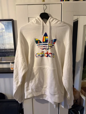 Vit hoodie från Adidas med färgglad logga - Vit hoodie från Adidas med stor, färgglad Trefoil-logga på bröstet. Klassisk känguruficka, snörning i huvan och mjukt material. Perfekt för dig som vill sticka ut med en unik och stilren look.