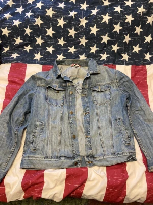 Ljusblå jeansjacka från Redwood Jeans - Snygg ljusblå jeansjacka från Redwood Jeans i klassisk modell med krage, bröstfickor med knappar och sidofickor. Jackan har metallknappar framtill och detaljerade sömmar. Perfekt för dig som gillar en avslappnad och cool stil. Storlek L. Jackan är i bra skick. 