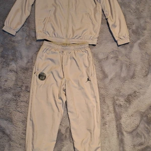 Beige PSG Jordan träningsset stl 147-158 - I fint skick. Sparsamt använt av ett barn. Köpt nytt från unisport. Säljer ett beige träningsset från PSG Jordan med dragkedja på jackan och elastisk midja på byxorna. Setet har PSG-logga, Jordan-logga och Qatar Airways-tryck. Perfekt för dig som gillar sportig och clean stil.