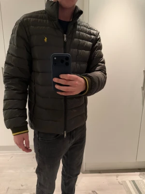 Mörkgrön pufferjacka från Polo Ralph Lauren - Snygg mörkgrön pufferjacka från Polo Ralph Lauren med gul broderad logga på bröstet. Jackan har ståkrage, dragkedja framtill och ribbade muddar med gul detalj. Perfekt för kyliga dagar och har en clean, sportig vibe.