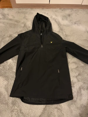 Svart vindjacka/regnjacka Lyle & Scott Junior - Svart vindjacka från Lyle & Scott Junior med huva och halv dragkedja framtill. Jackan har två sidofickor med dragkedja och en gul logga på bröstet. Perfekt för blåsiga och regniga dagar och har en clean, sportig look.