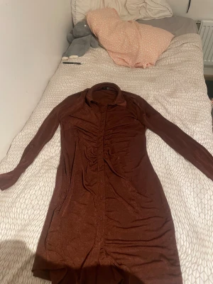 Brun långärmad klänning Vero Moda - Snygg brun långärmad klänning från Vero Moda med krage och knappar framtill. Klänningen har rynkade detaljer längs mitten och är i ett mjukt, stretchigt material som sitter skönt på kroppen. Perfekt för dig som gillar en stilren look med twist. Är också glimmrig 