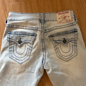 True Religion Billy Relaxed Bootcut Jeans - Ljusblå jeans från True Religion, modell Billy Relaxed Bootcut. Snygga kontrastsömmar i blått, klassiska ficklock med knappar och ikonisk logga på bakfickorna. Lös Bootcut passform. Nypris runt 2300. Lite lite slitna längst ner men inget man tänker på! Det är en herrmodell men passar för kvinnor med!