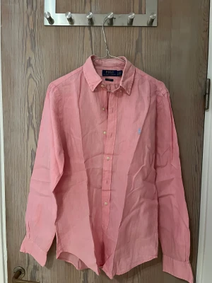 Rosa linneskjorta från Polo Ralph Lauren - Snygg rosa skjorta från Polo Ralph Lauren i luftig linnematerial. Klassisk krage, långa ärmar och knappar framtill. Diskret broderad logga på bröstet i ljusblått. Perfekt för dig som gillar stilrena och fräscha plagg med en chill vibe.