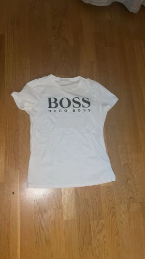 Vit t-shirt från Hugo Boss - Snygg vit t-shirt från Hugo Boss med stor svart logga på bröstet. Klassisk rund hals och korta ärmar. Tillverkad i mjuk bomull för skön känsla. Perfekt för dig som gillar stilrena och enkla plagg med tydlig branding.