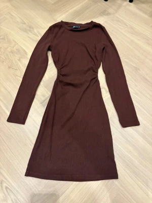 Brun ribbad långärmad klänning Zara - Säljer en brun ribbad klänning från Zara i storlek M. Modellen är figurnära med lång ärm och har snygg rynkdetalj i midjan som ger en cool siluett. Materialet är mjukt och stretchigt, perfekt för dig som gillar enkel men stilren look.