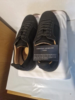 Svarta sneakers från Christian Louboutin - Snygga svarta sneakers från Christian Louboutin med lyxig mocka och klassisk röd sula. Skorna har snörning, rund tå och diskret broderad logga på hälen. Insidan är beige och sulan är platt. Perfekt för dig som vill sticka ut med exklusiv stil.