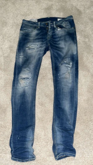 Blå Dondup jeans - Säljer ett par blå dondup jeans med modellen George och slitna detaljer på benen. Jeansen har klassisk femficksdesign, dragkedja och knappstängning. Färgen är mörkblå med ljusare tvätt och små färgstänk för en cool, använd look.