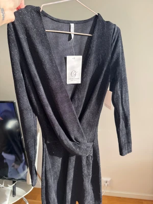 Omlottklänning Chiara Forthi - Supersnygg svart omlottklänning från Chiara Forthi med glittrigt tyg och v-ringning. Klänningen har långa ärmar och markerad midja, perfekt för dig som vill sticka ut. Sitter tajt och har en elegant look med twistad detalj framtill. Aldrig använd mer än testad, prislapp kvar.