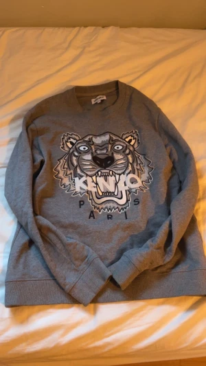 Grå Kenzo sweatshirt med tigertryck - Säljer en grå sweatshirt från Kenzo med det ikoniska tigerhuvudet och texten 'Kenzo Paris' på bröstet. Tröjan har rund hals, långa ärmar och ribbade muddar. Perfekt för dig som gillar streetstyle och vill sticka ut med ett coolt print passar ungefär vid längd 180..