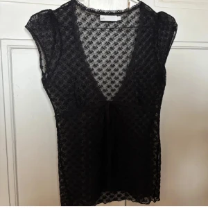 Svart spetsblus med knyt från att. - Svart transparent spetsblus från att. med blommigt mönster, djup v-ringning och knyt framtill. 