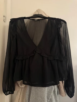 Svart volangblus från Bik Bok - Säljer en svart transparent blus från Bik Bok med långa ärmar och djup v-ringning. Blusen har volangdetaljer längs axlar och midja.
