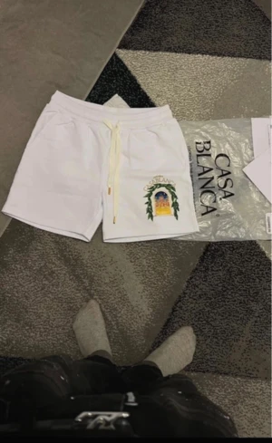 Vita shorts från Casablanca med tryck - Säljer ett par fräscha vita shorts från Casablanca med snyggt färgglatt tryck på ena benet och gul snörning i midjan. De har en relaxed fit och är perfekta för sommaren. Materialet känns mjukt och skönt, 100% bomull.