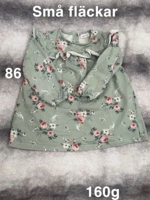 Grön blommig klänning med volang - Supersöt grön klänning från  Newbie med blommigt mönster i rosa och vitt. Klänningen har långa ärmar, volangdetalj över bröstet och knäppning bak i nacken. Perfekt för dig som gillar gulliga och bekväma outfits!🌸 Små fläckar. Storlek 86