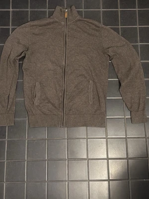 Montego Stickad Cardigan / Full-Zip – Brun – Strl S - Kan gå ner i pris ordentligt vid snabb affär! Den har hel dragkedja framtill och en hög krage som värmer gott. Tröjan är utrustad med två praktiska fickor på sidorna samt ribbade muddar vid både ärmslut och nederkant för en bra passform. Materialet känns mjukt och den är perfekt att ha som ett värmande lager över en t-shirt eller skjorta. 