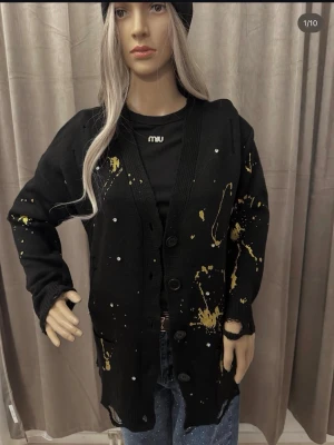 Svart kofta med guldstänk och pärldetaljer - Svart stickad kofta med knappar framtill, dekorerad med guldiga färgstänk och små pärlliknande detaljer. Koftan har lång ärm och en lite oversized passform. Perfekt för dig som gillar en edgy och unik stil.