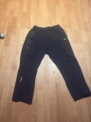 Svarta NOCTA mjukisbyxor från Nike - Svarta mjukisbyxor från Nike NOCTA med gul logga och detaljer. Byxorna har dragsko i midjan, raka ben och tryckta detaljer på både fram- och baksida. Tillverkade i mjukt material som passar perfekt för chill eller träning. Endast använd 4 gånger!