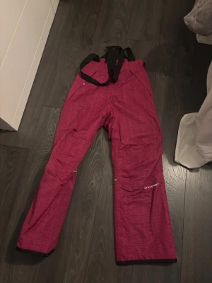 Röda täckbyxor Everest strl 158 - Täckbyxor från Everest i rosa färg med diskret rutigt mönster. Byxorna har svarta justerbara hängslen och reflexdetaljer på benen. Perfekta för vinterns alla äventyr och håller dig varm och torr i backen.
