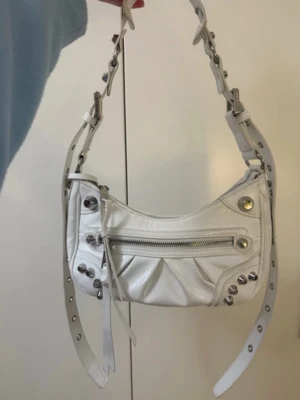 Vit axelväska från Steve Madden - Säljer en snygg vit axelväska från Steve Madden i skinnimitation. Väskan har coola silverdetaljer, dragkedja framtill och justerbart axelband med nitar. Medföljer matchande liten plånbok och rund spegel. Perfekt för dig som gillar edgy accessoarer.