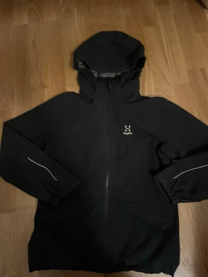 Svart vindjacka från Haglöfs Junior - Säljer en svart vindjacka från Haglöfs i juniorstorlek 156. Jackan har huva, hel dragkedja framtill, två fickor och reflekterande detaljer på ärmarna. Insidan är fodrad med mesh för extra komfort. Perfekt för blåsiga dagar!