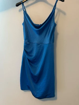 Blå kort klänning med tunna band - Säljer en blå kort klänning med tunna axelband och draperad front. Klänningen har en mjuk, glansig yta och dragkedja i ryggen. Perfekt för dig som vill ha en enkel men snygg look.