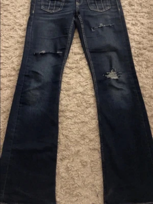 Lågmidjade bootcut jeans - Säljer ett par mörkblå bootcut jeans från Gina Tricot i storlek 158. Jeansen har slitna detaljer och hål på benen för en trendig look. Klassisk femficksmodell med vita kontrastsömmar och knapp med logga. 
